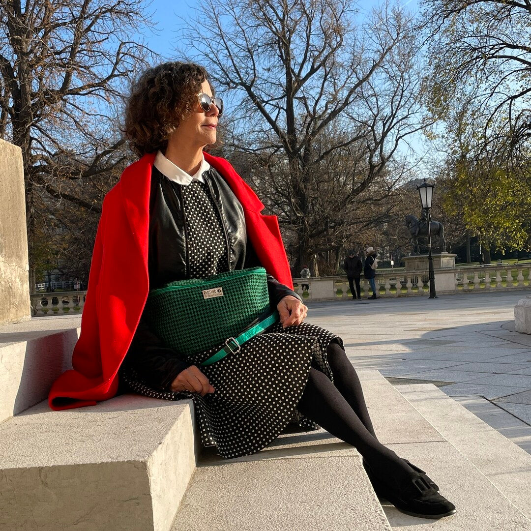 Frau in getupftem Kleid und roter Jacke sitzt mit grüner Bum Bag aus TWEED Stoff auf Stufen im Park
