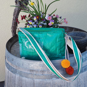 grüne Umhängetasche mit Bommeln steht auf einem Fass neben Blumen