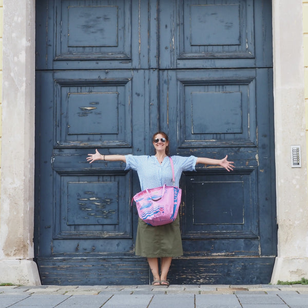 Frau steht mit rosa Badetasche vor riesiger blauer Tür und lacht
