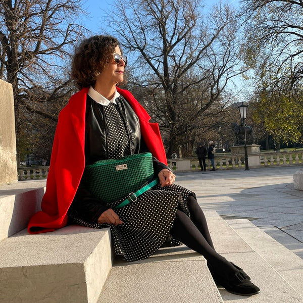 Frau in getupftem Kleid und roter Jacke sitzt mit grüner Bum Bag aus TWEED Stoff auf Stufen im Park