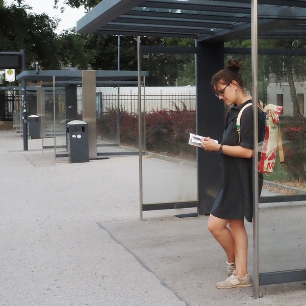 Frau steht an der Bushaltestelle, trägt beige farbenen Rucksack und liest ein Buch